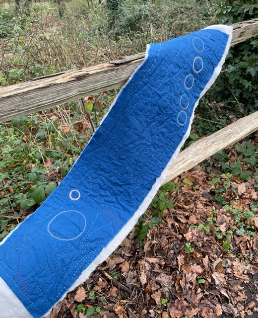 A strip of dark blue fabric, quilted with a design of circles, curves and lines. The piece is backed with grey linen, and rests on a wooden fence in woodland.