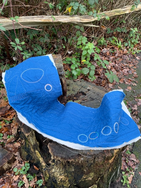 A strip of dark blue fabric, quilted with a design of circles, curves and lines. The piece is backed with grey linen, and rests on a tree stump on a woodland path