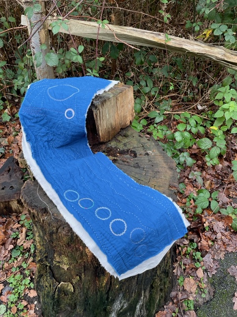 A strip of dark blue fabric, quilted with a design of circles, curves and lines. The piece is backed with grey linen, and rests on a tree stump
