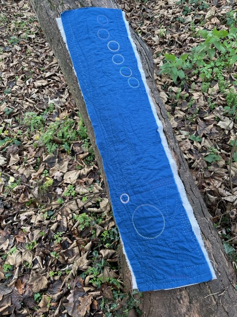 A strip of dark blue fabric, quilted with a design of circles, curves and lines. The piece is backed with grey linen, and rests on log against a carpet of leaves..